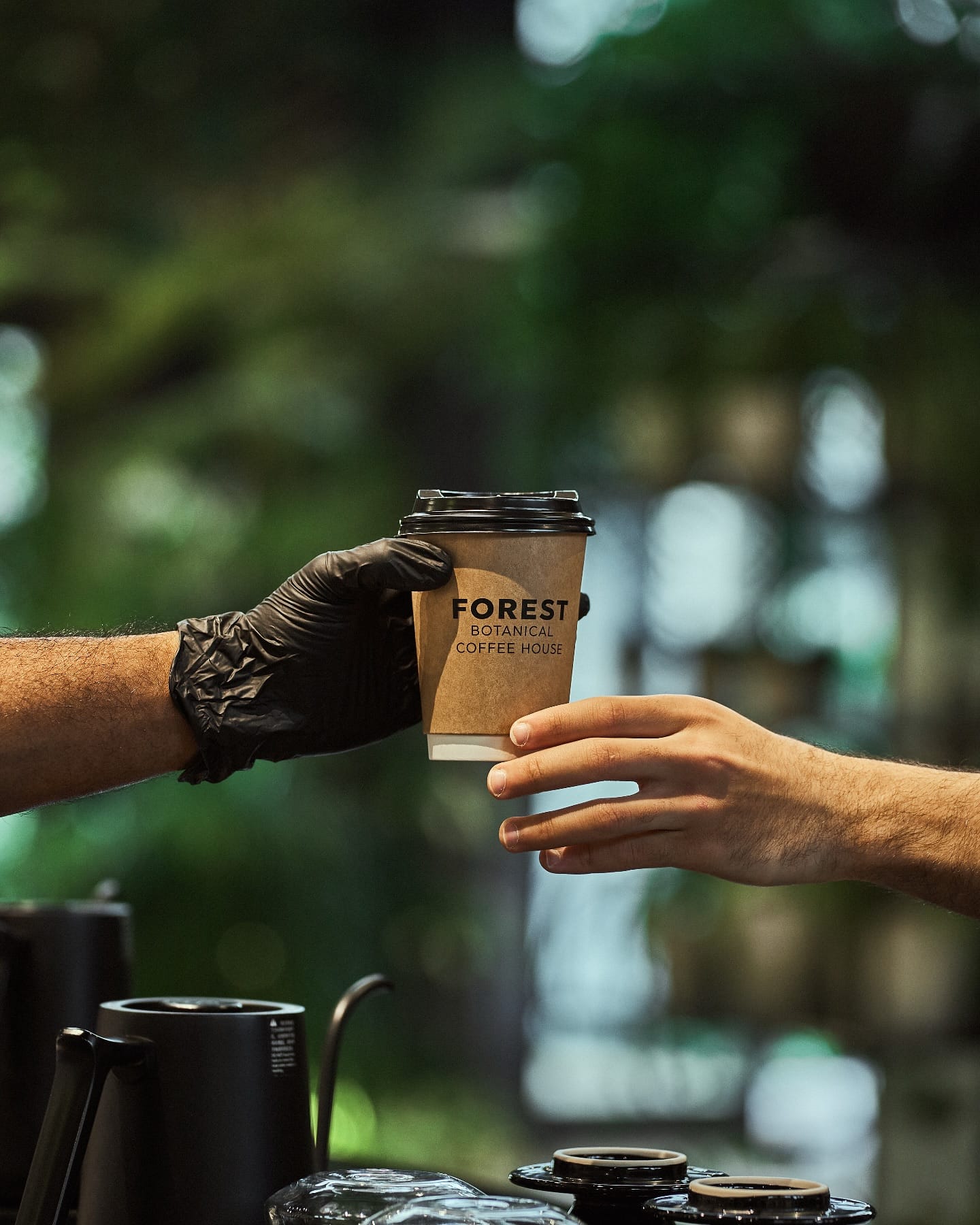 A barista's black-gloved hand passing a Forest takeaway cup to a guest, a gooseneck kettle and drippers in front
